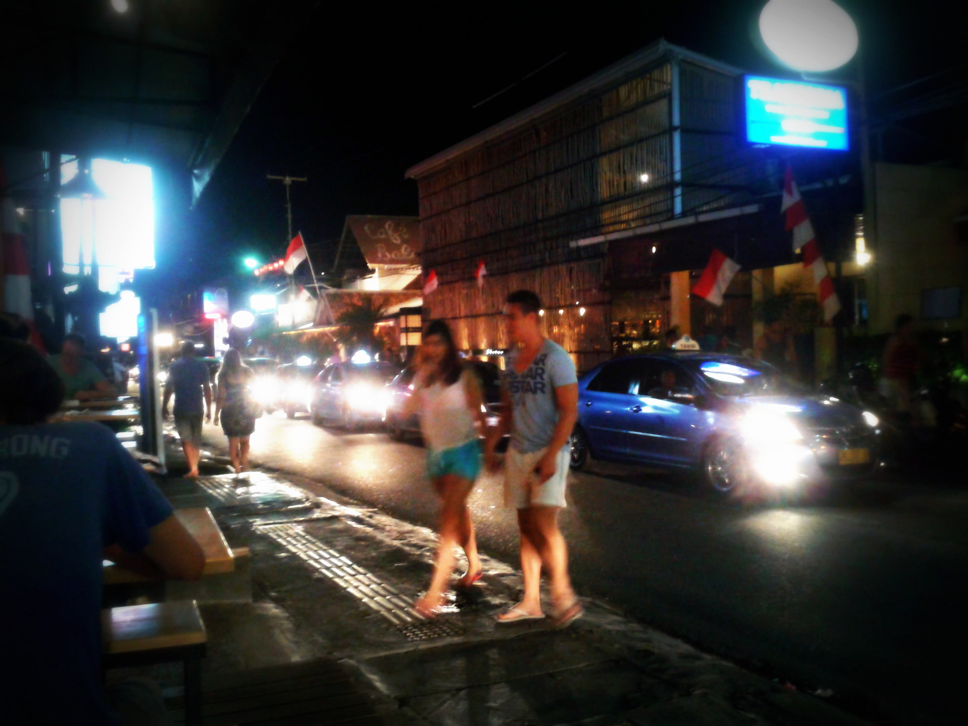 People crossing a lit-up Bali street at night between cars and scooters