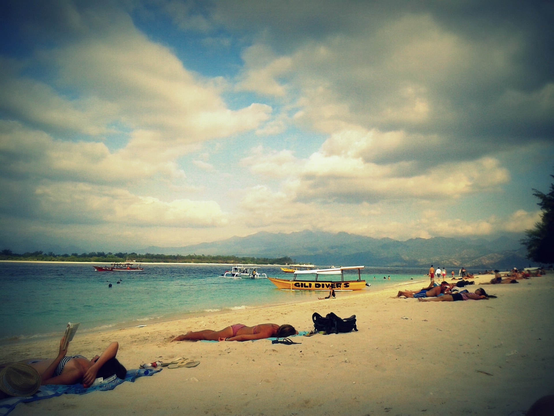 A yellow Gili Divers boat anchored off the beach with sunbathers on the sand and Lombok's mountains behind