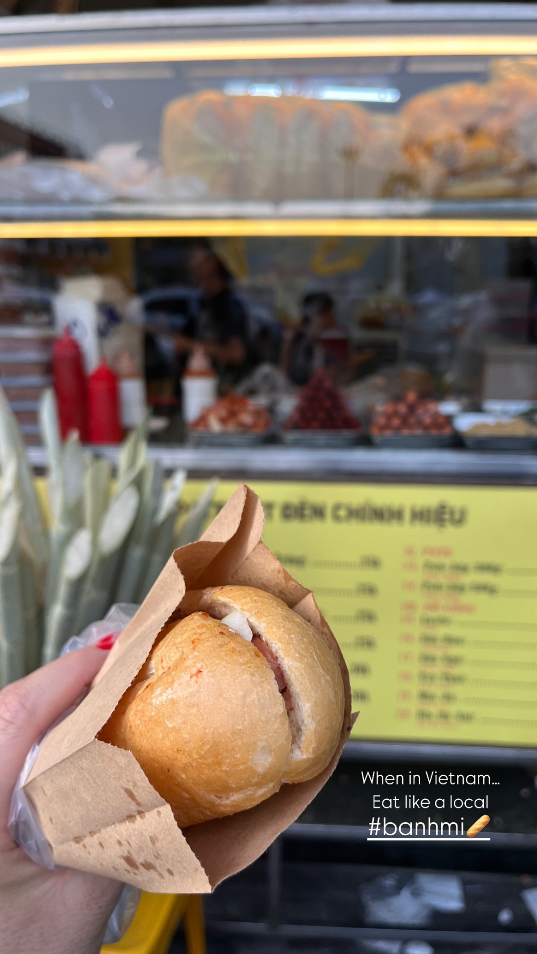 A banh mi sandwich in paper held in front of a Hanoi street food stall
