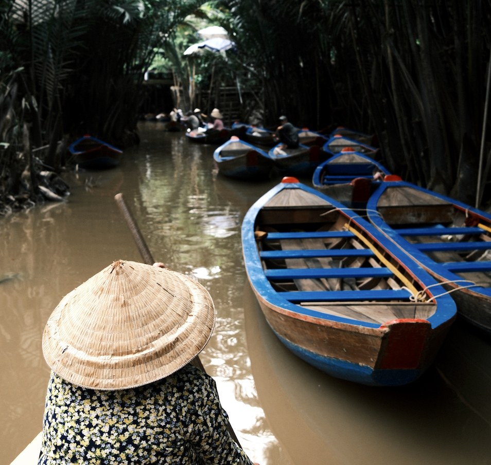 A woman in a conical hat rowing a wooden boat through a Mekong Delta palm canal
