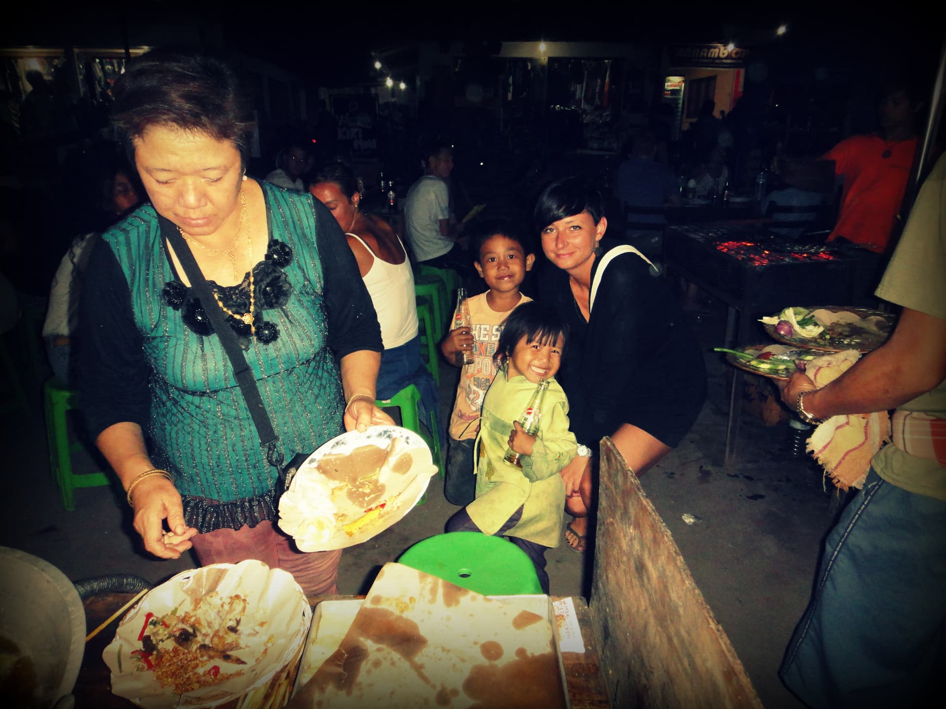 A traveller posing with two local children at a busy Bali night market food stall at night