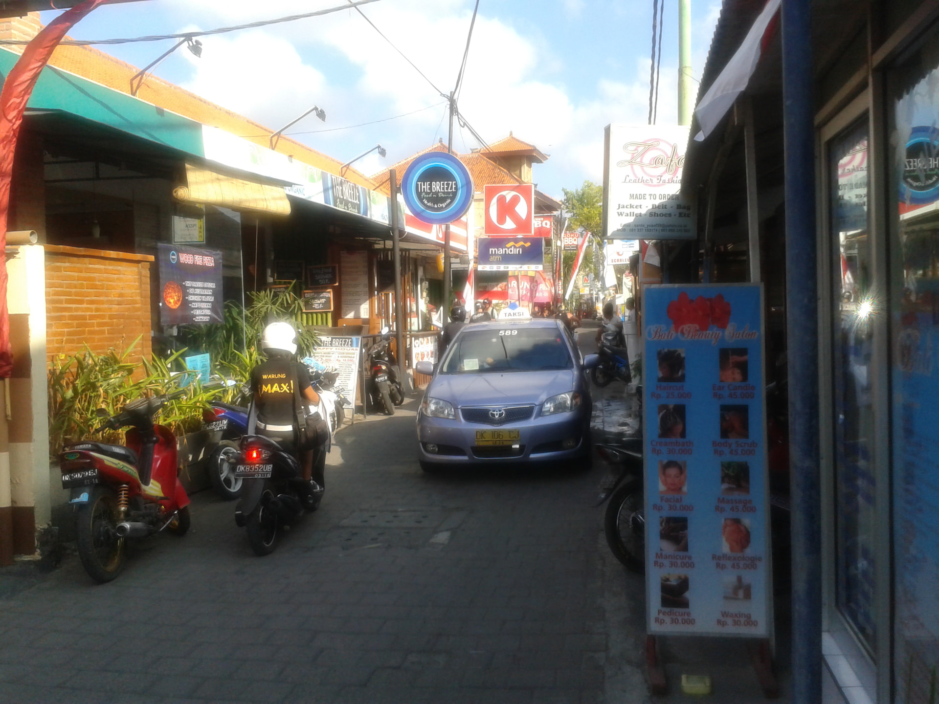 A narrow Kuta side street with parked scooters, warung signs and a taxi squeezing through