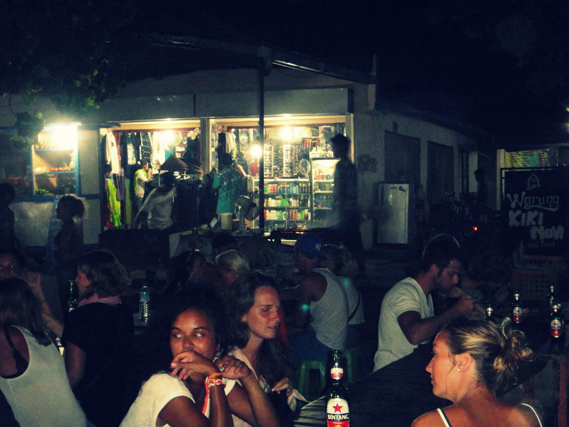Travellers eating dinner at long tables beside a lit-up warung and night market stalls in Bali