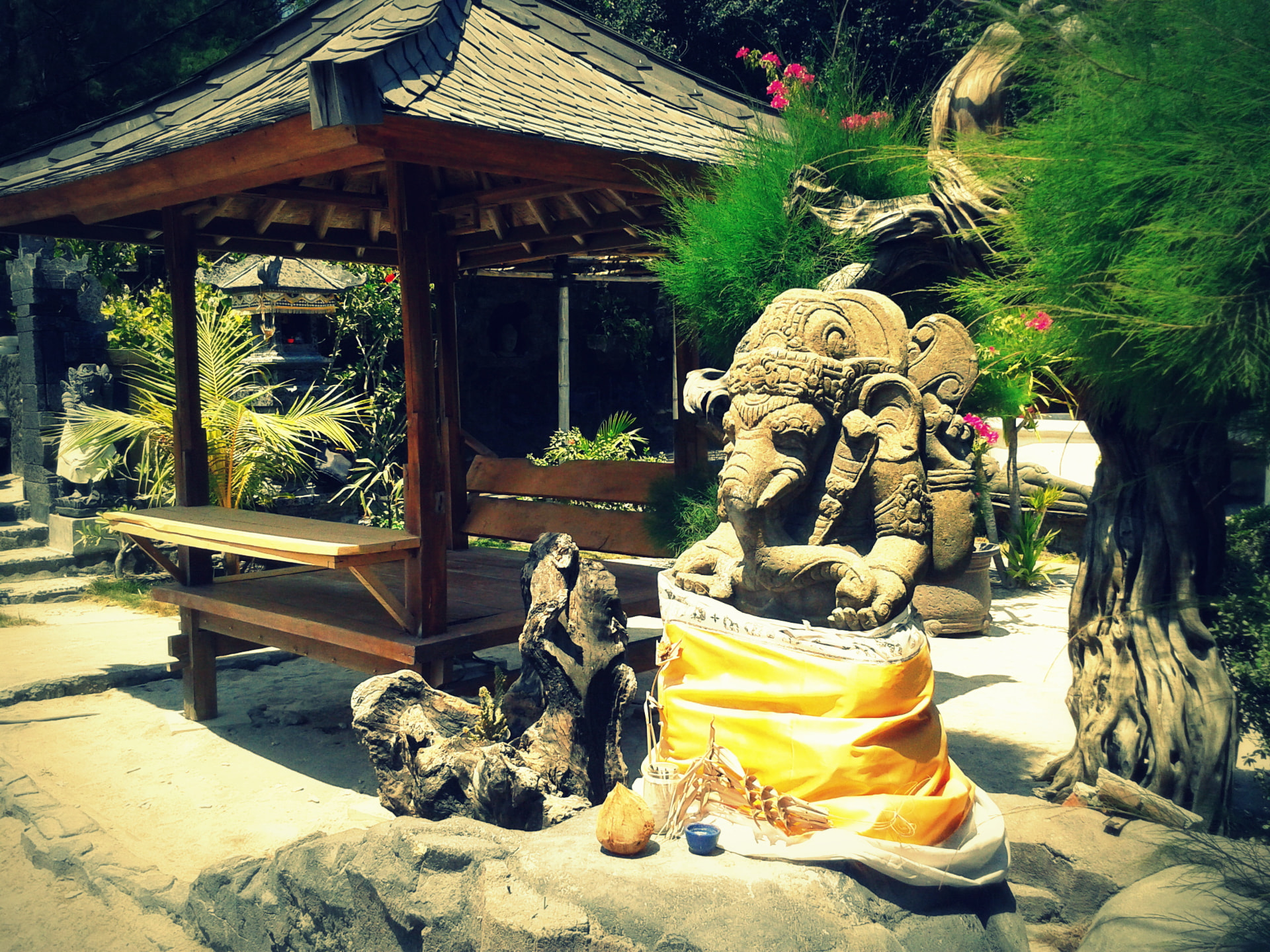 A stone Ganesha statue wrapped in yellow cloth with offerings at its base beside a wooden garden pavilion in Bali