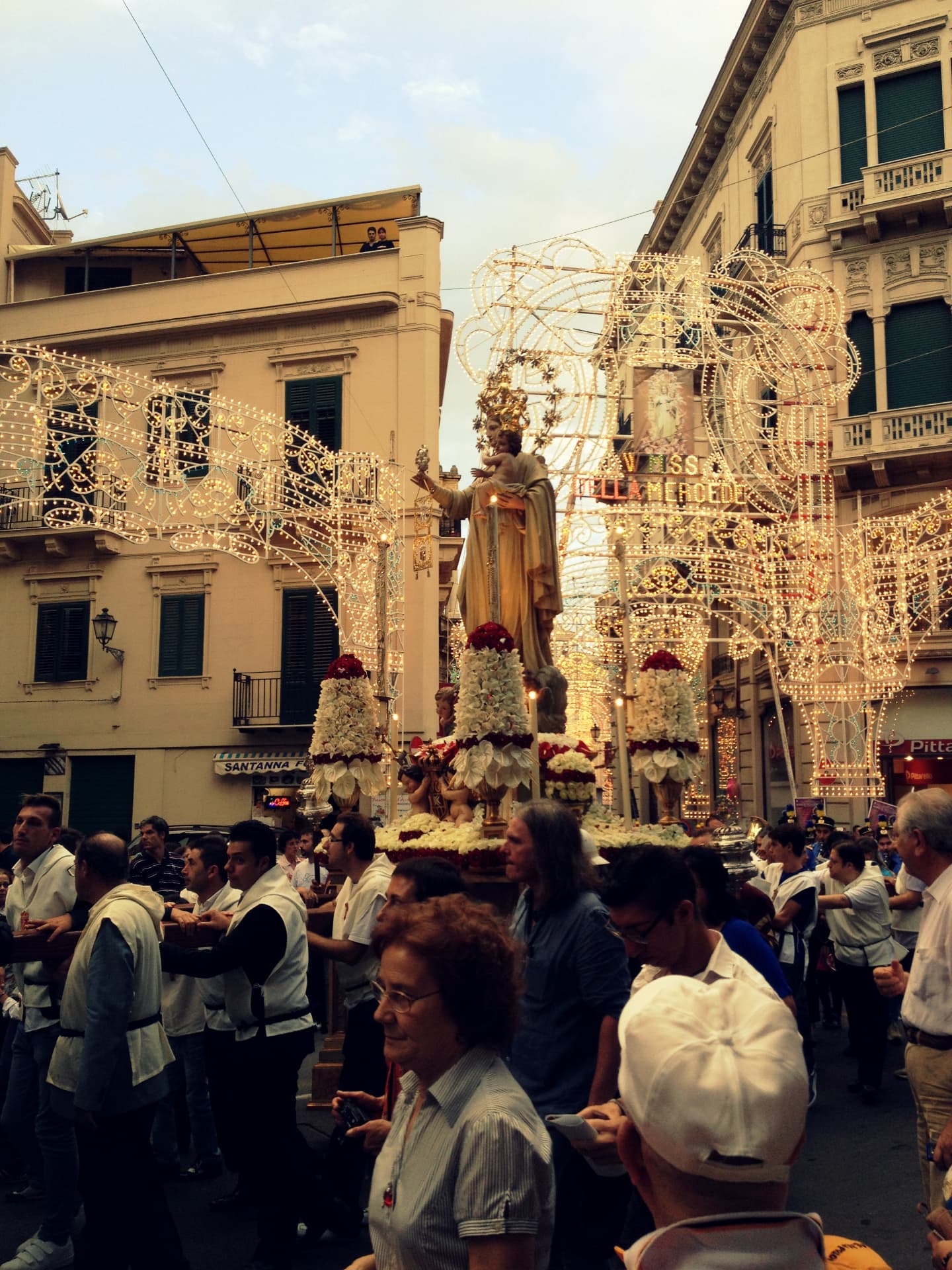 A night procession moving beneath elaborate strings of festival lights in Palermo