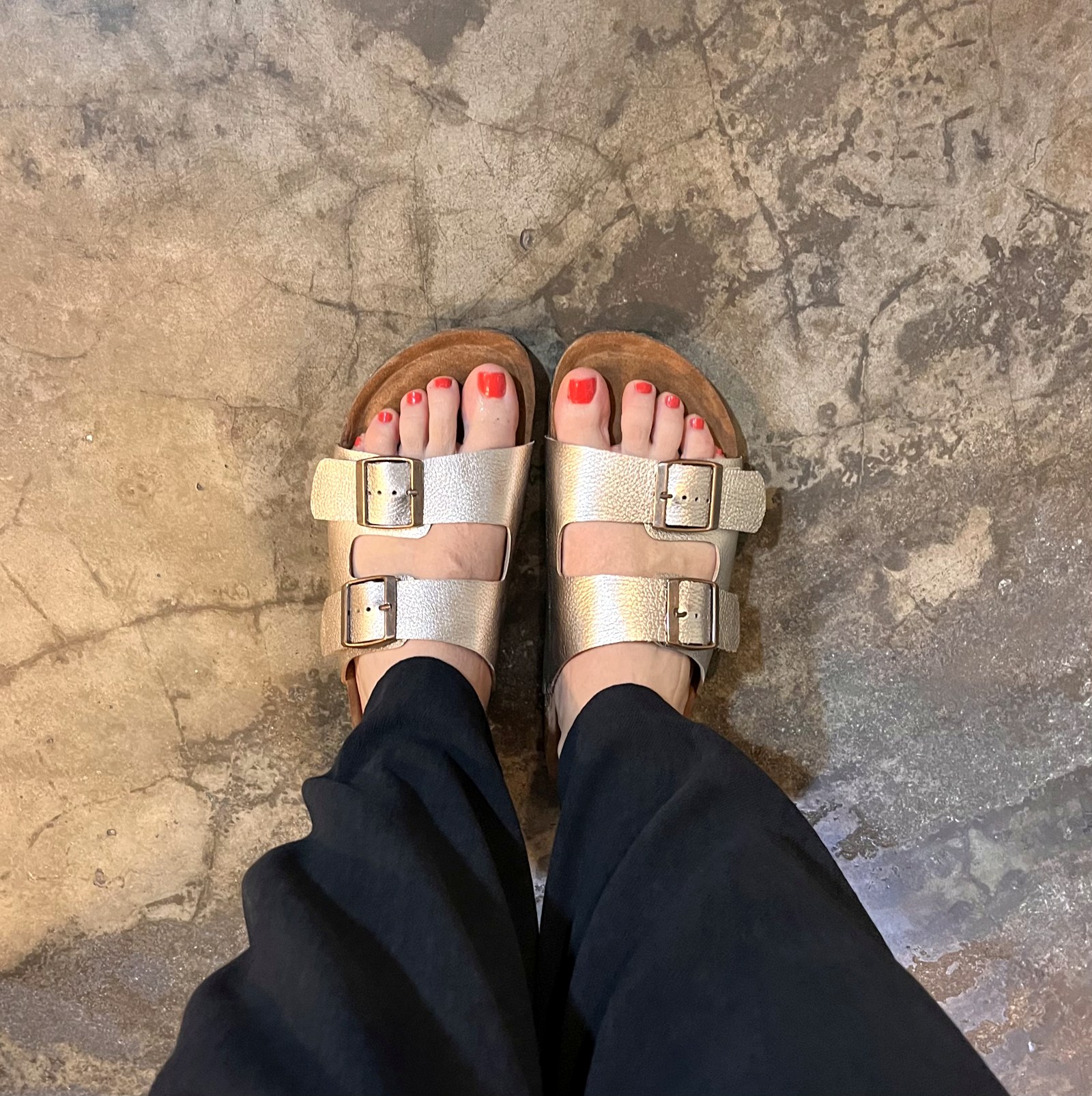 Andreja's feet in gold buckle sandals with red painted toenails, seen from above on a stone floor