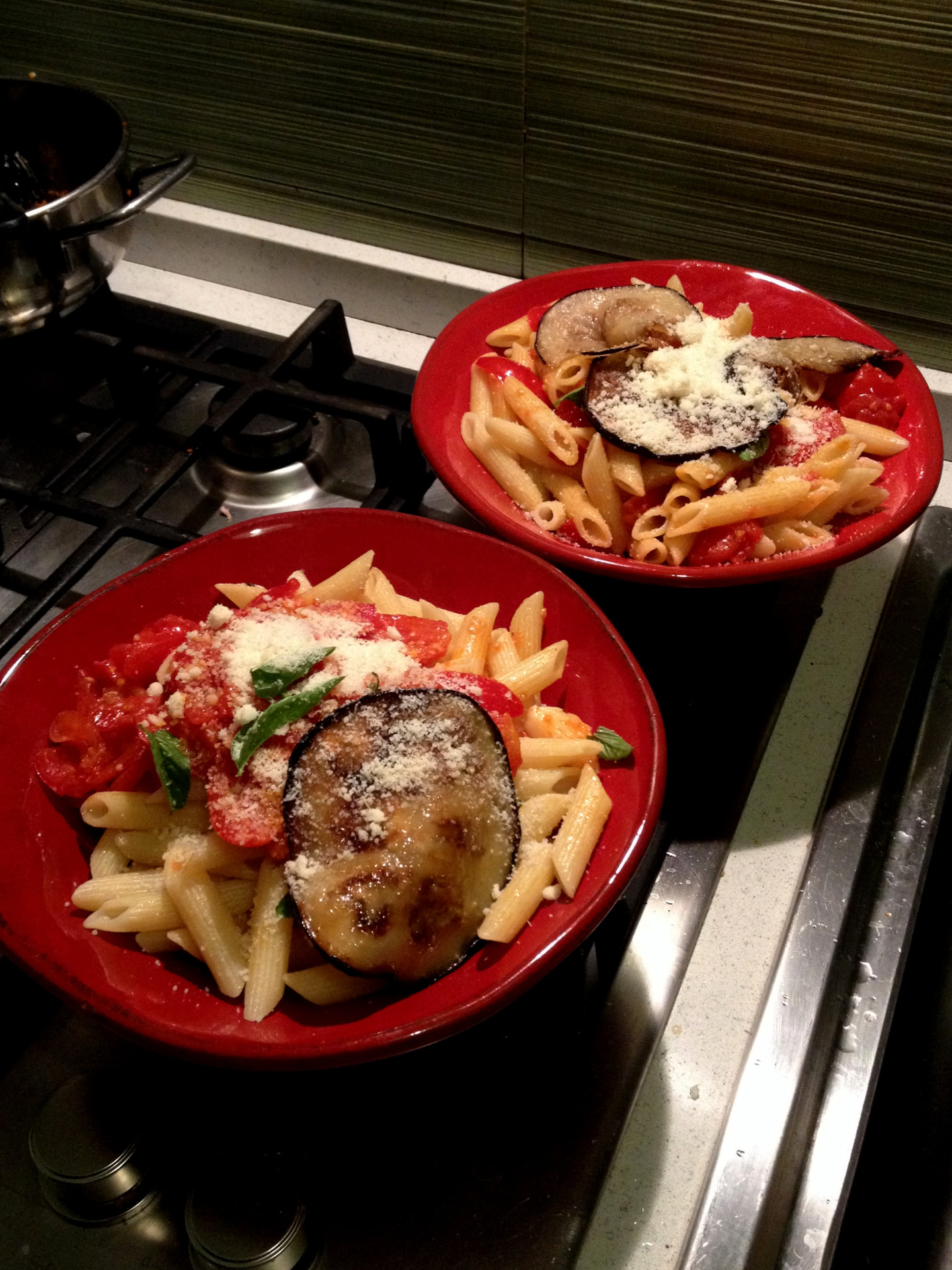 Two red bowls of pasta alla norma with aubergine and ricotta salata on a kitchen stove