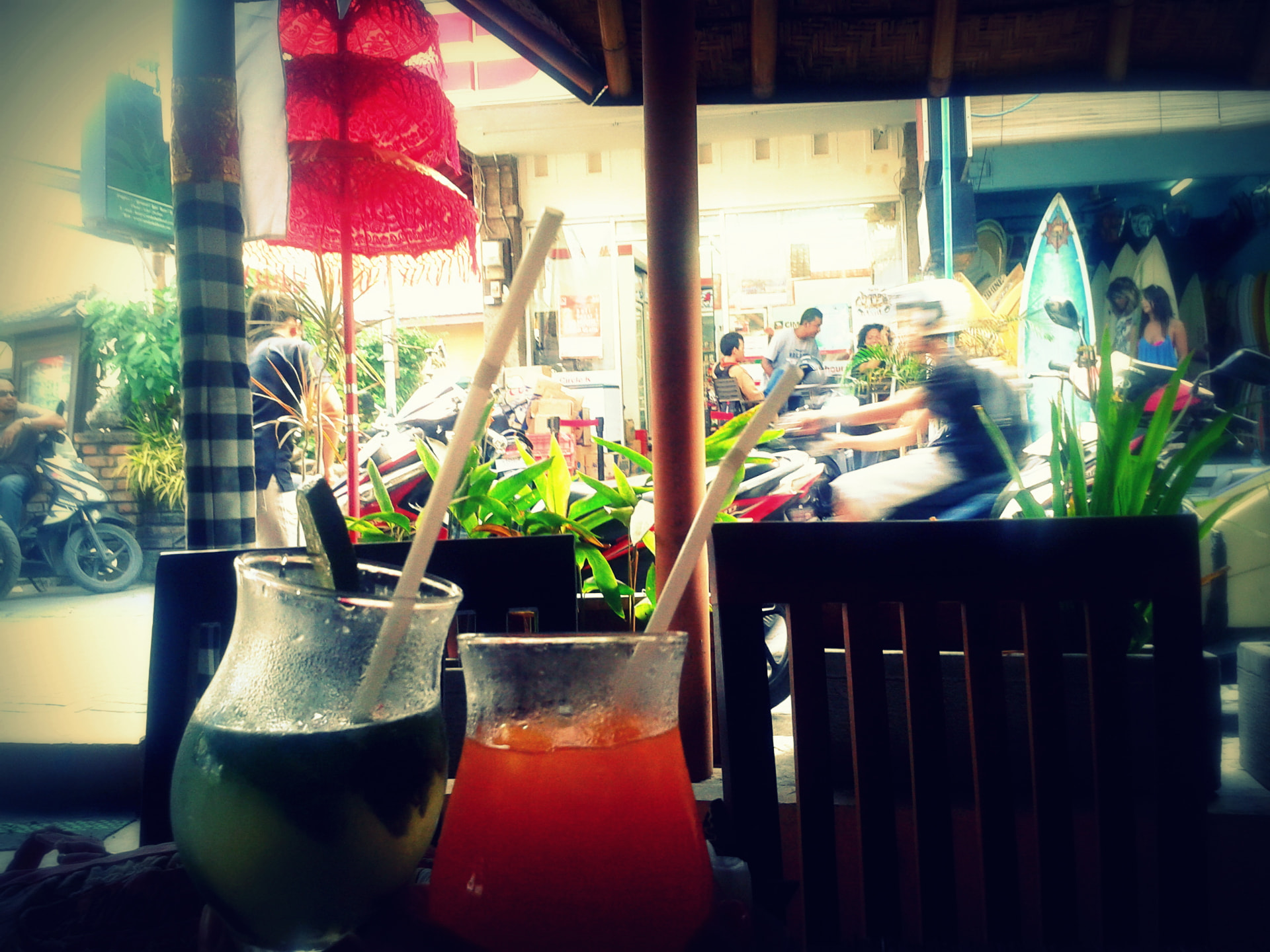 Two cold cocktails with straws on a café table in Kuta, scooters and a surf shop blurred behind