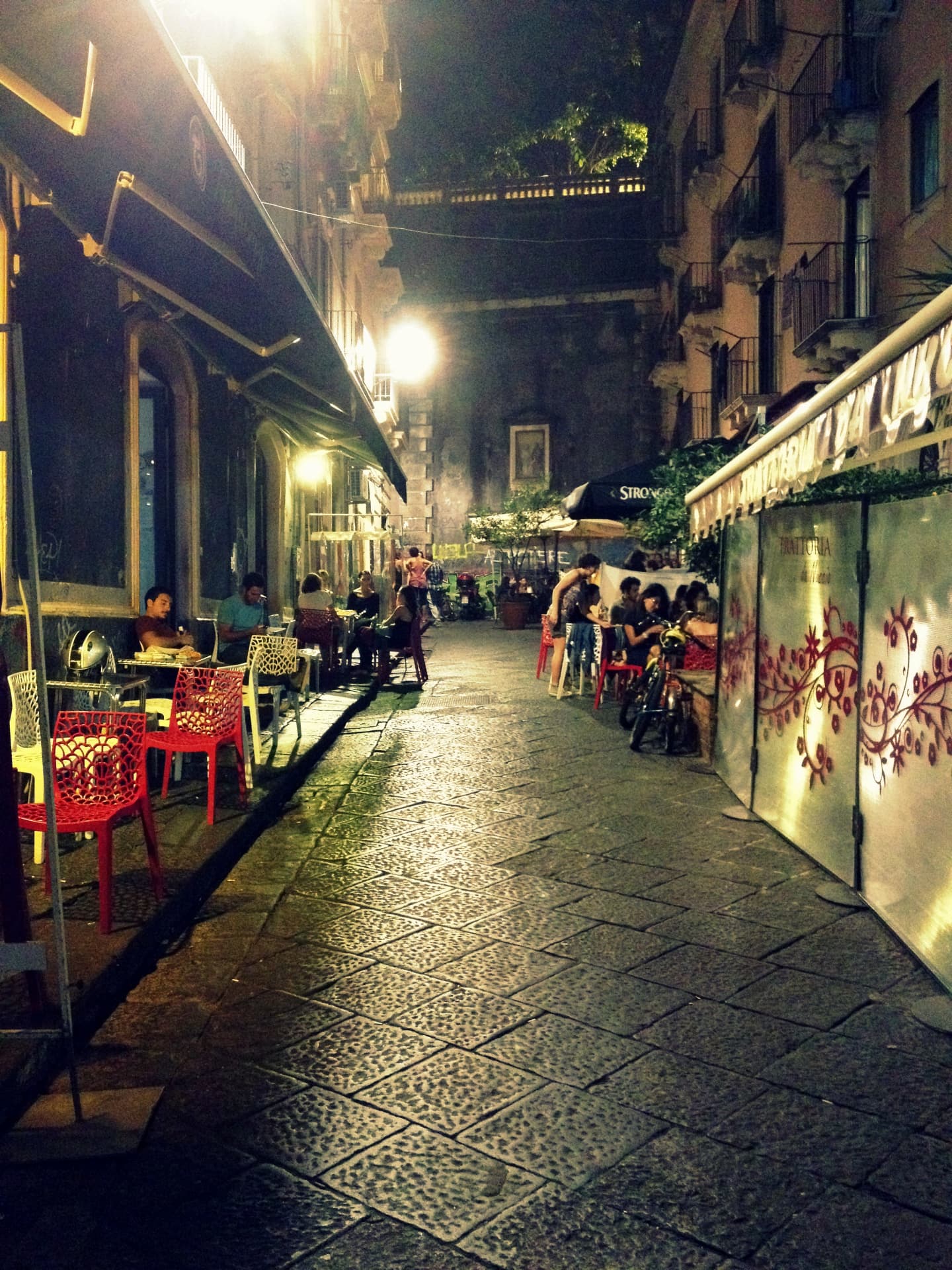 A narrow Catania alley at night with red chairs, lit trattoria awnings and people eating outside