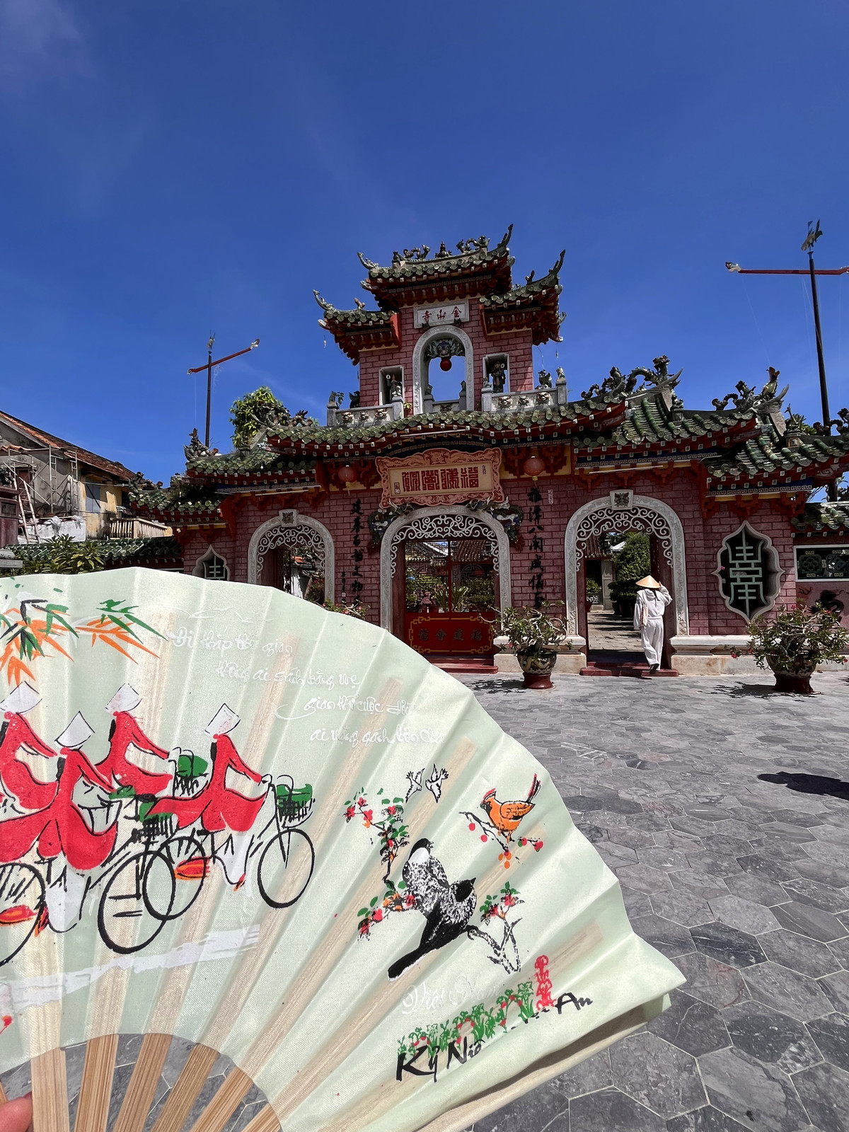 Hand-painted Hoi An fan held up in front of the ornate Fujian Assembly Hall gate under a blue sky