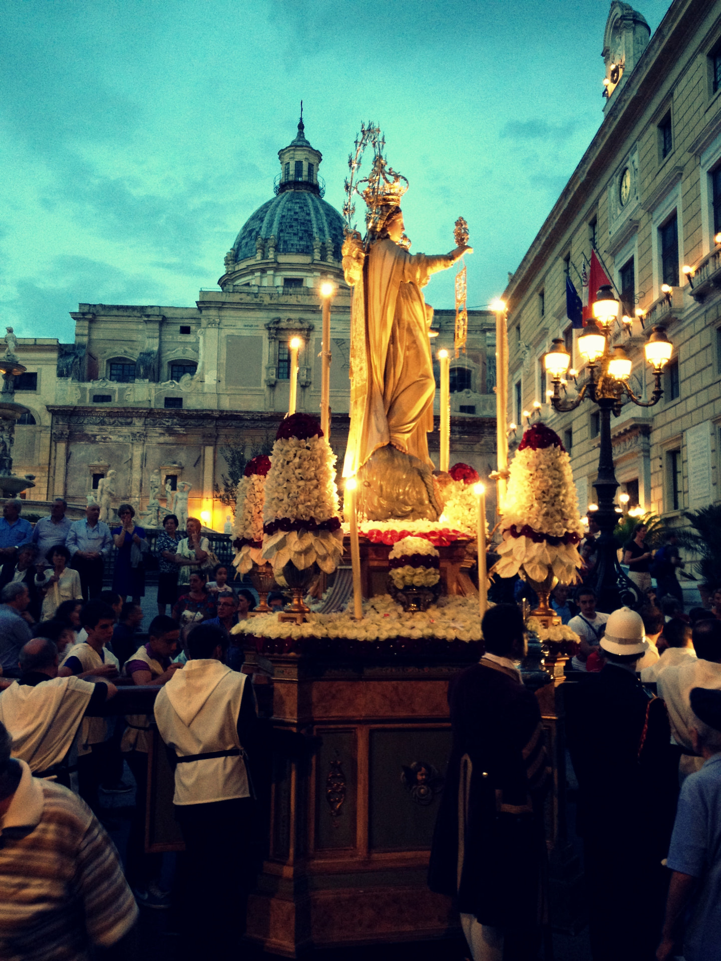 A gilded statue of the Madonna carried on a flower-covered float at dusk in Palermo