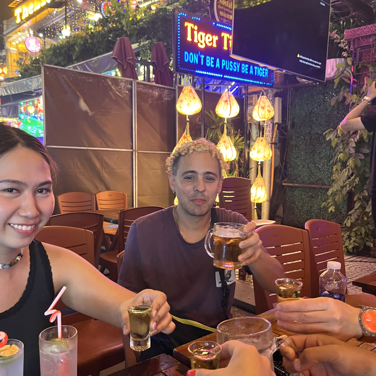 Friends raising shot glasses under the neon Tiger bar sign reading Don't be a pussy, be a tiger in Hoi An
