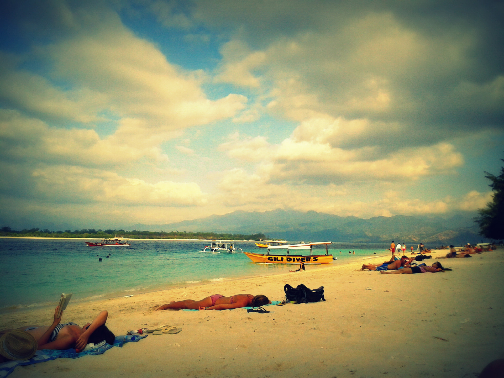 A yellow Gili Divers boat anchored off the beach with sunbathers on the sand and Lombok's mountains behind