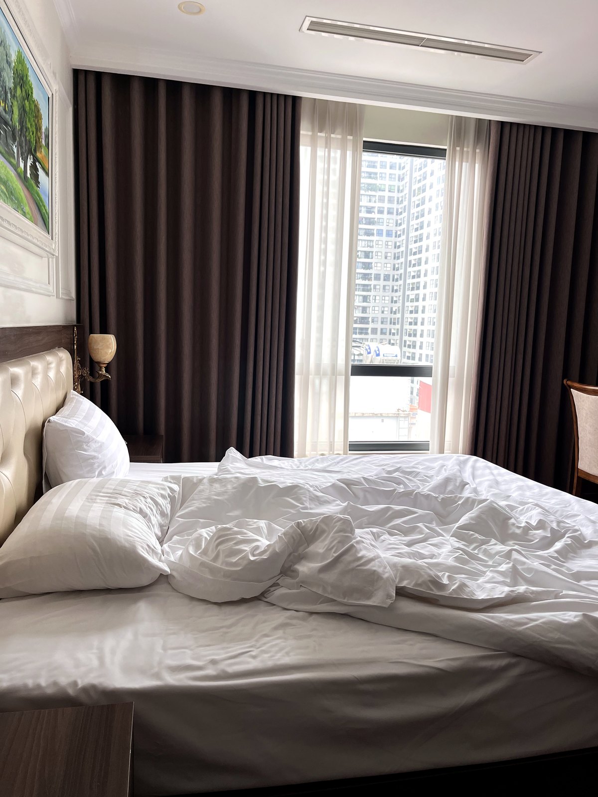 Unmade white hotel bed by a tall window with dark curtains in Hanoi