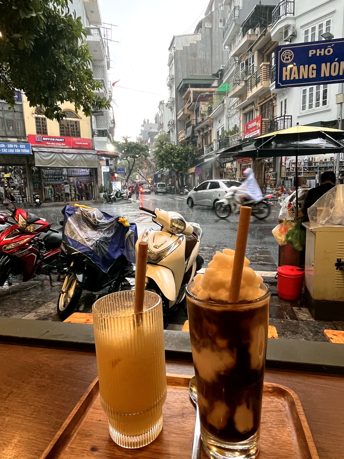 Pineapple juice and iced coffee on a cafe counter overlooking a rainy Hanoi street with parked scooters