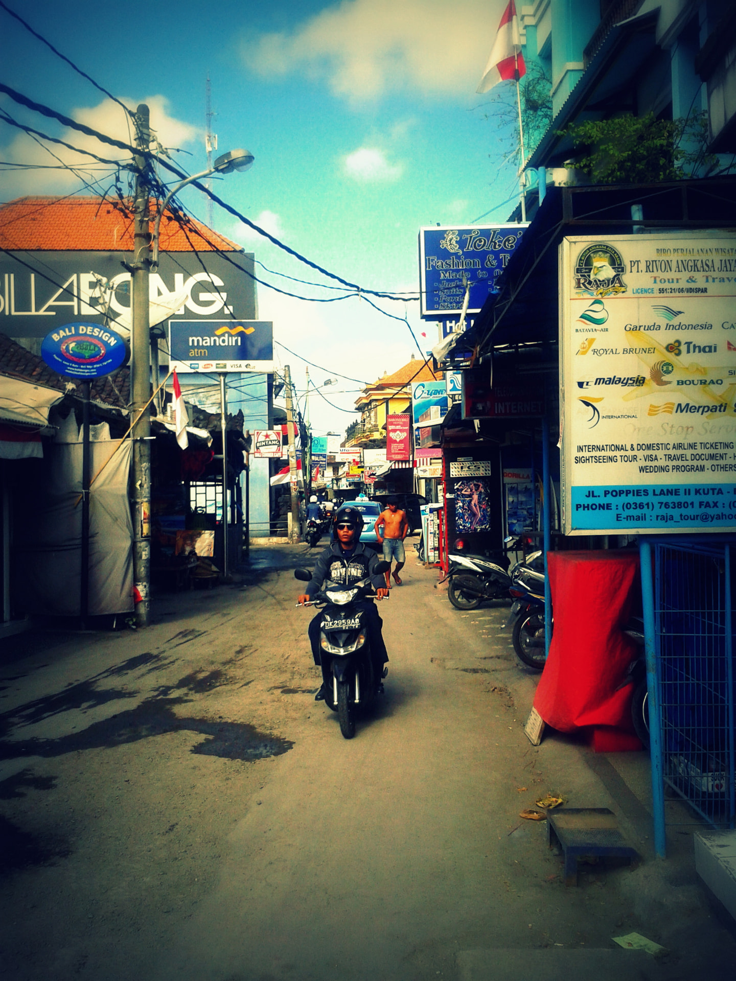 A rider on a scooter coming down a narrow lane in Kuta lined with shops and travel agencies
