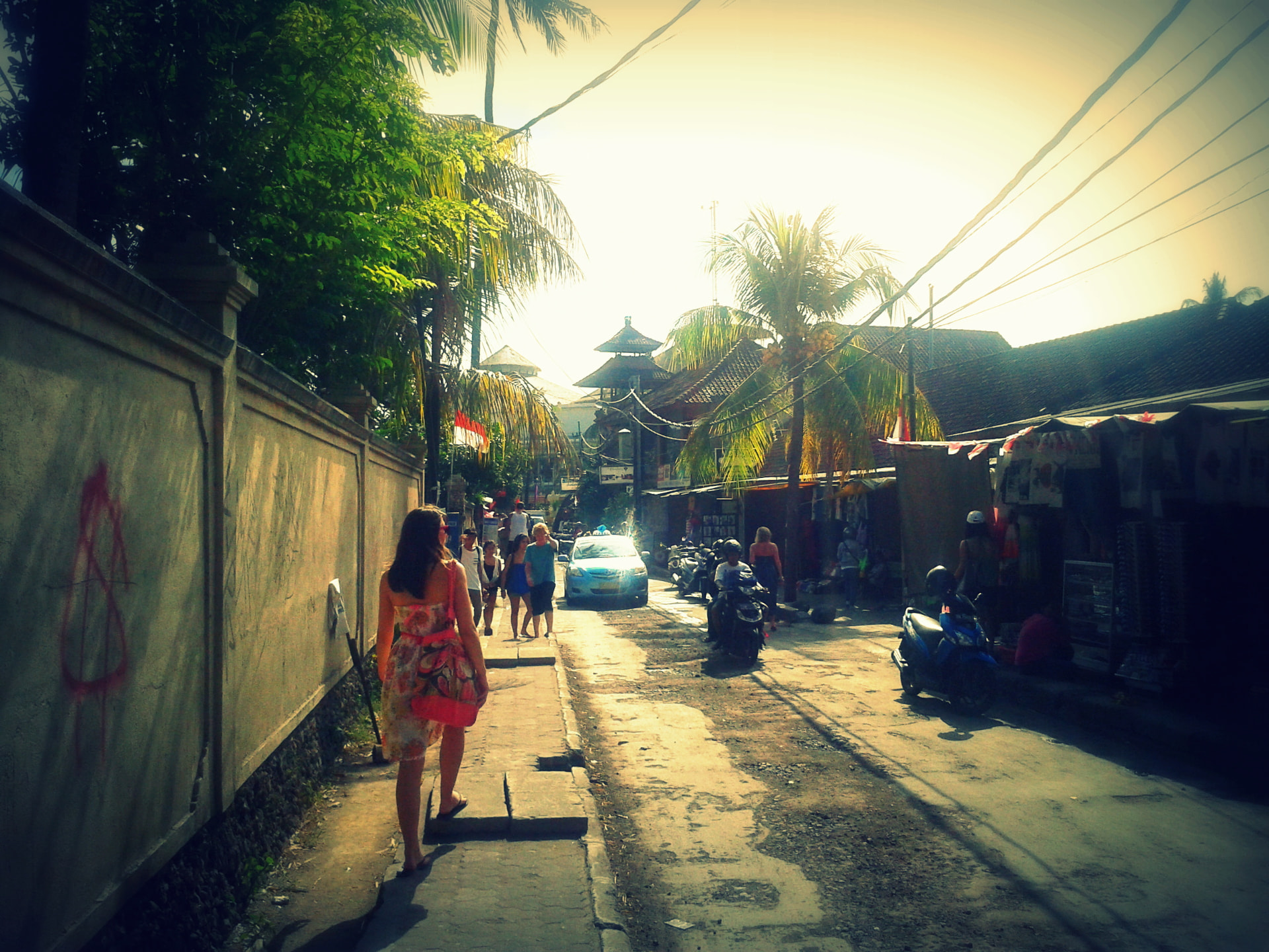 A woman in a floral dress walking down a sunlit Bali street lined with palms and parked scooters
