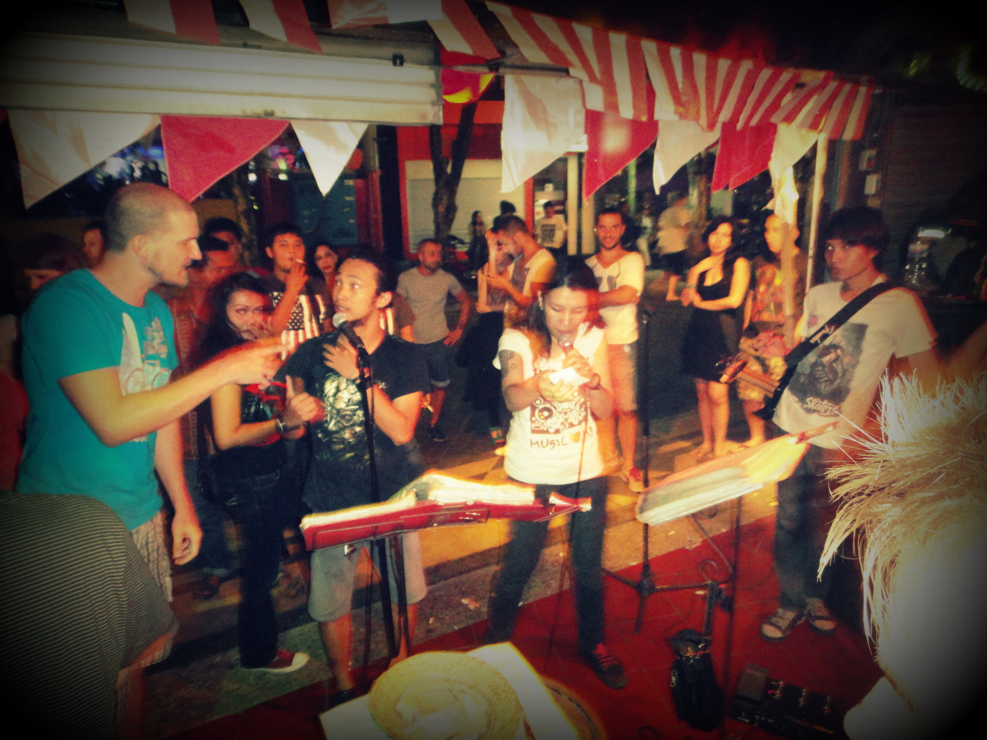 A crowd of locals and travellers singing street karaoke under red and white bunting at night in Kuta, Bali