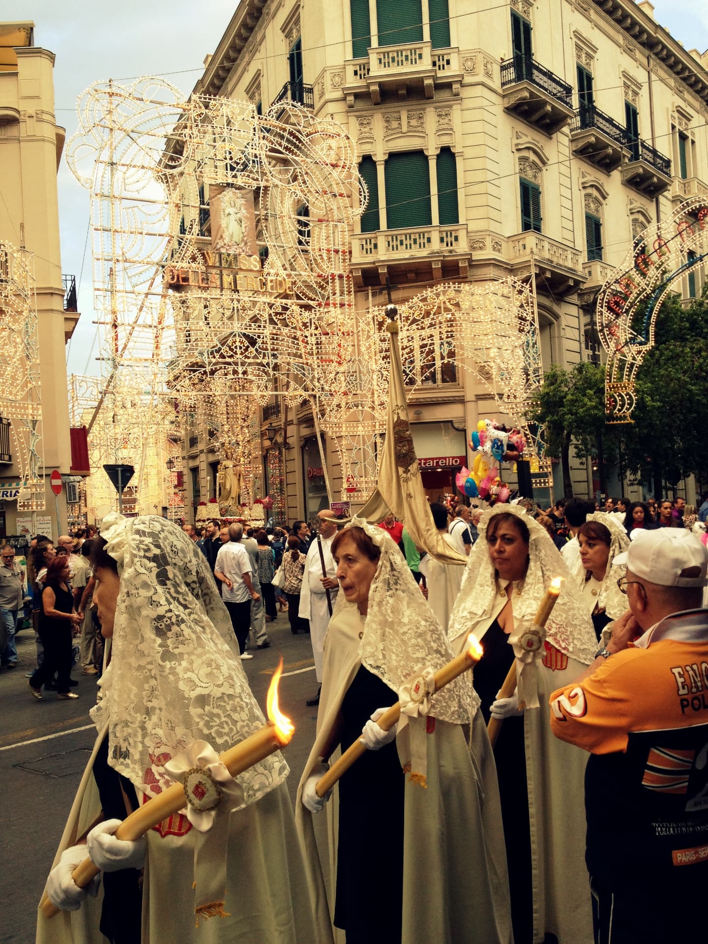 A religious procession with lace veils and candles under illuminated street arches in Palermo