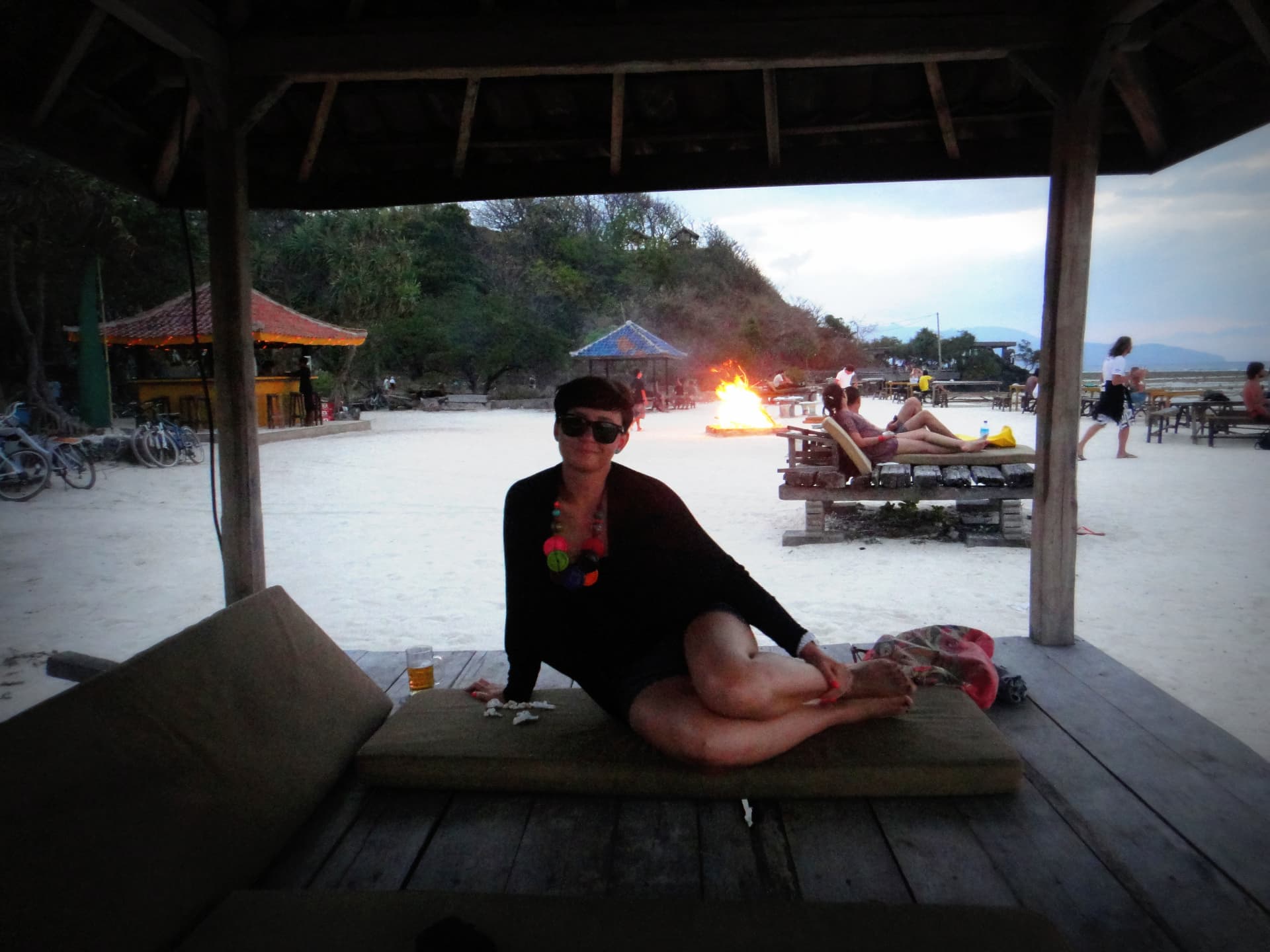A woman relaxing on a beach lounge at dusk with a bonfire glowing on Gili Trawangan