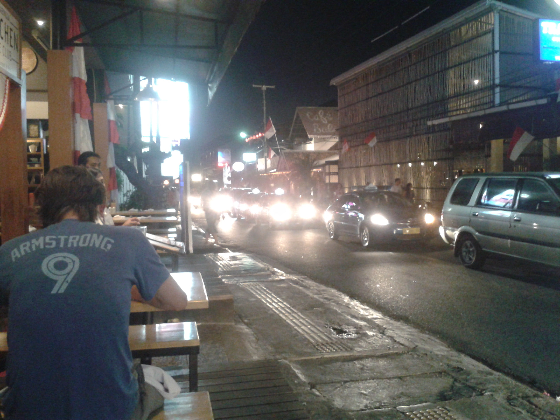 A busy Bali street at night seen from a roadside table, car headlights and lit-up cafés
