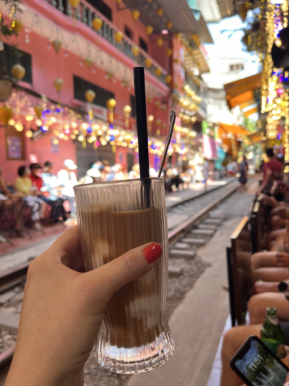 Iced Vietnamese coffee held up in front of Hanoi's train street lined with pink buildings and lanterns