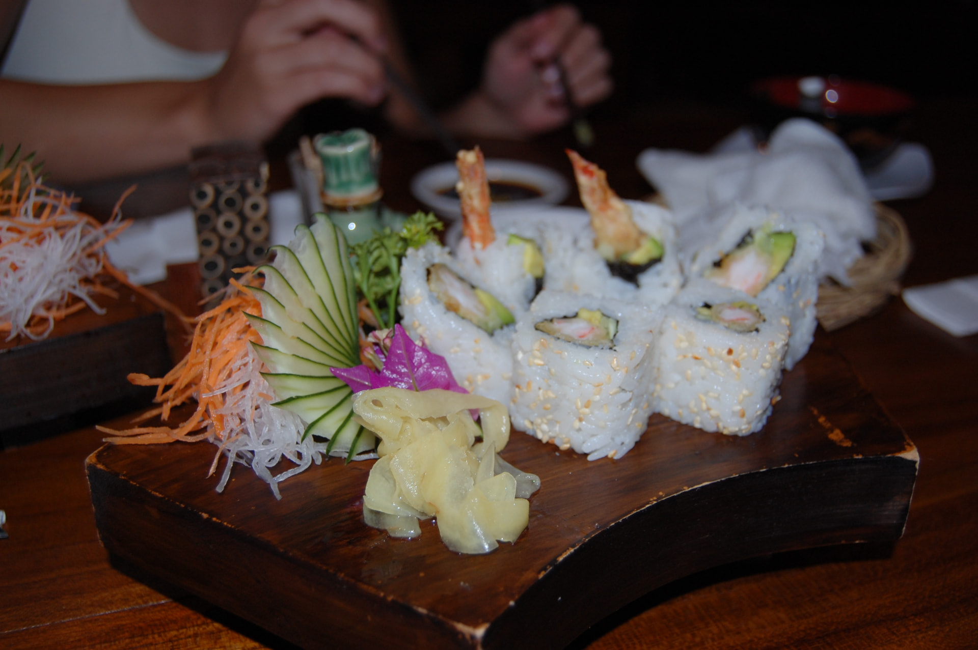 A sushi platter with tempura prawn rolls, pickled ginger and carved cucumber on a wooden board at a dark restaurant in Kuta