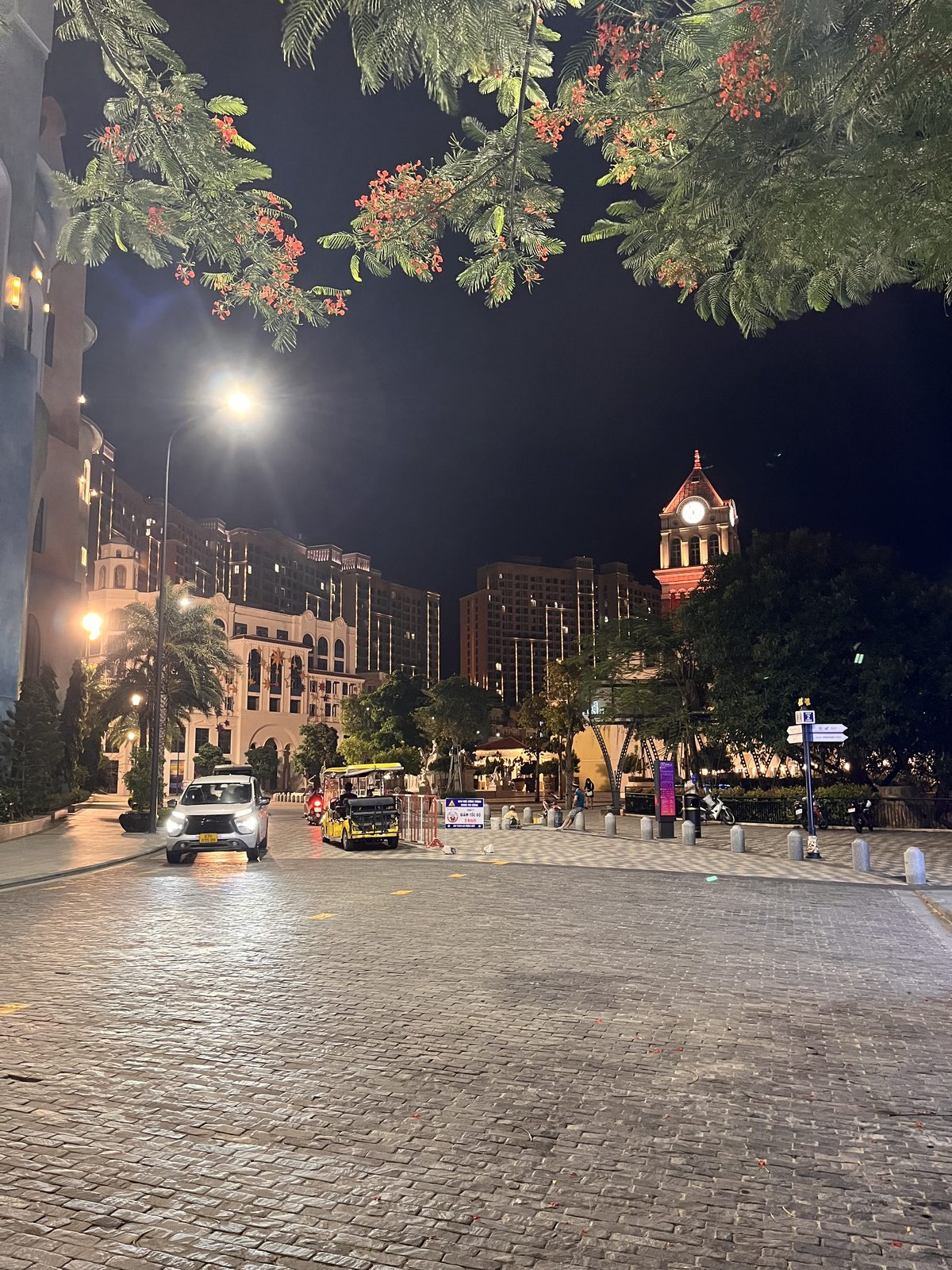 Night street in Sunset Town on Phu Quoc with an illuminated clock tower, blossoming trees and a yellow shuttle