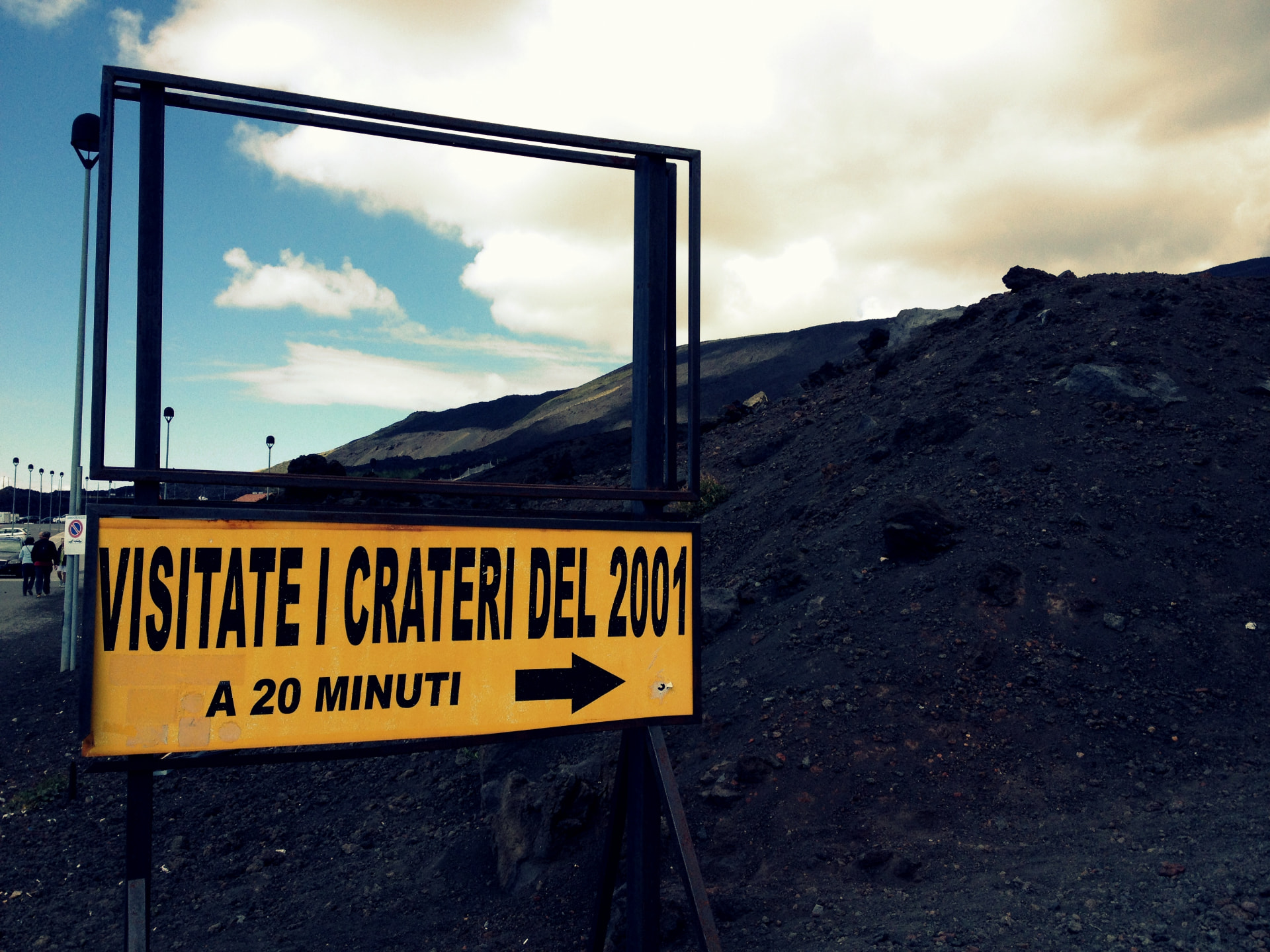 A yellow sign on Mount Etna reading Visitate i crateri del 2001, a 20 minuti, beside black volcanic slopes