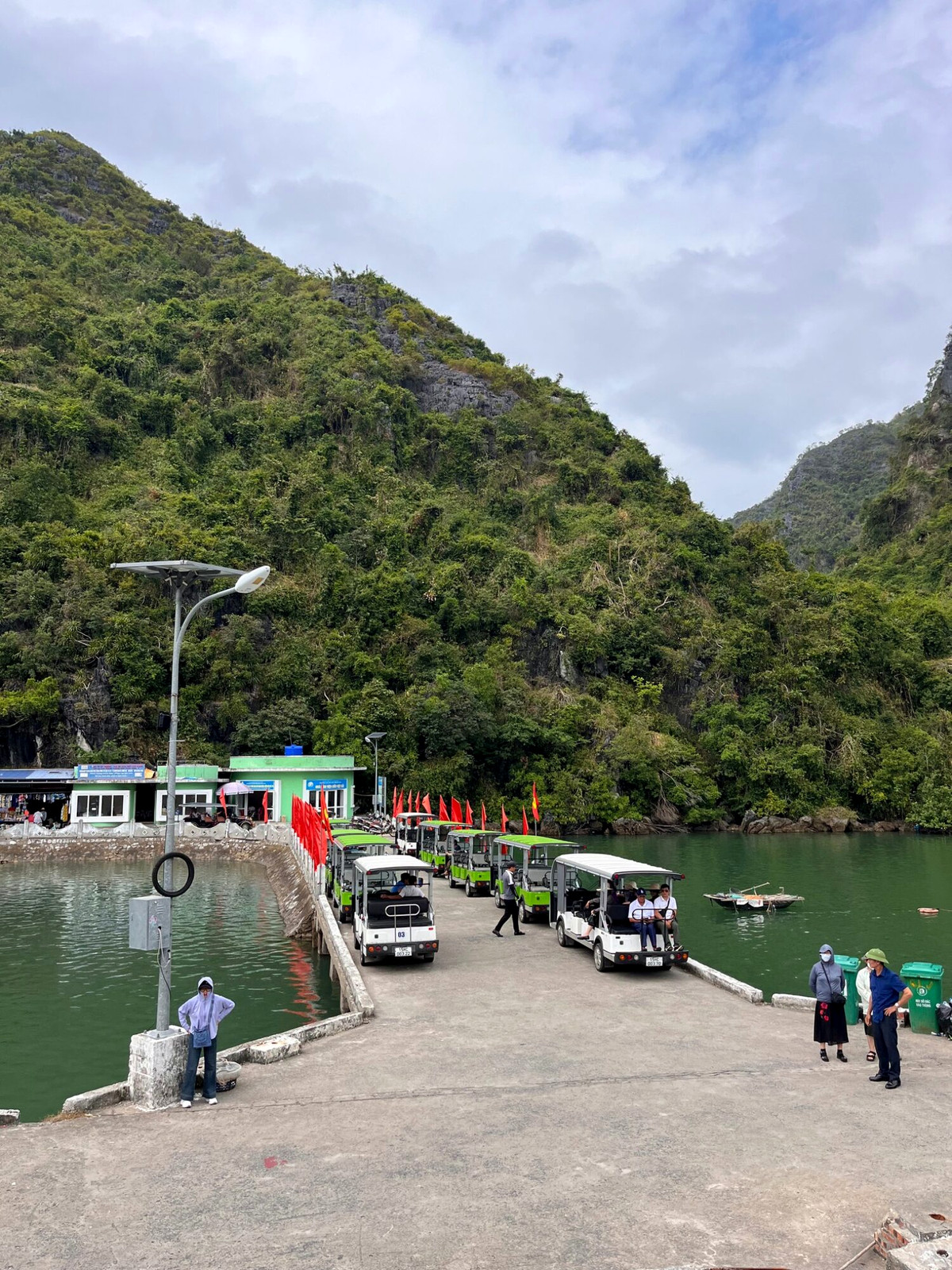 People and parked vehicles at a village stop beneath wooded Cat Ba hills