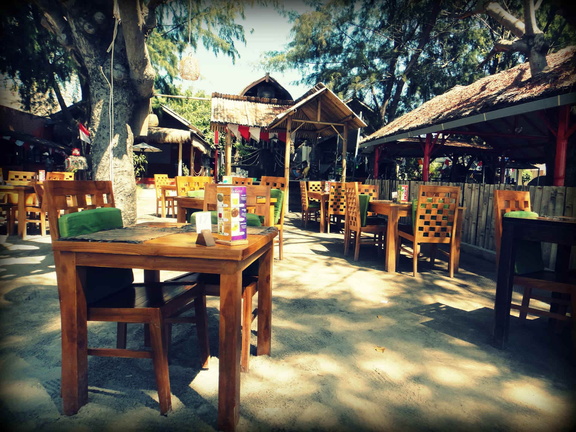 Empty wooden tables under trees at a sandy beachside breakfast spot in Bali