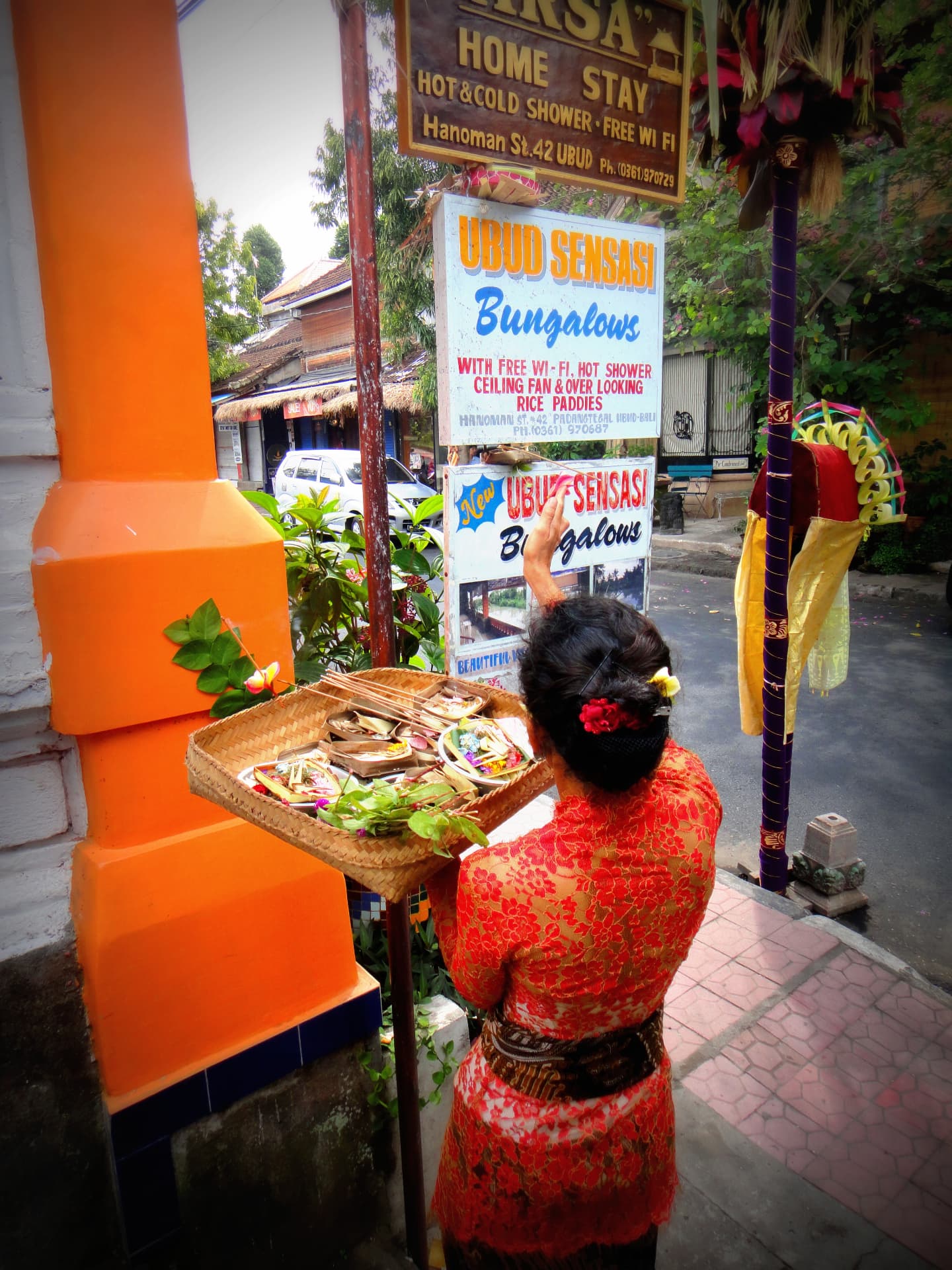 A Balinese woman in a red kebaya placing a tray of canang sari offerings on a street shrine in Ubud