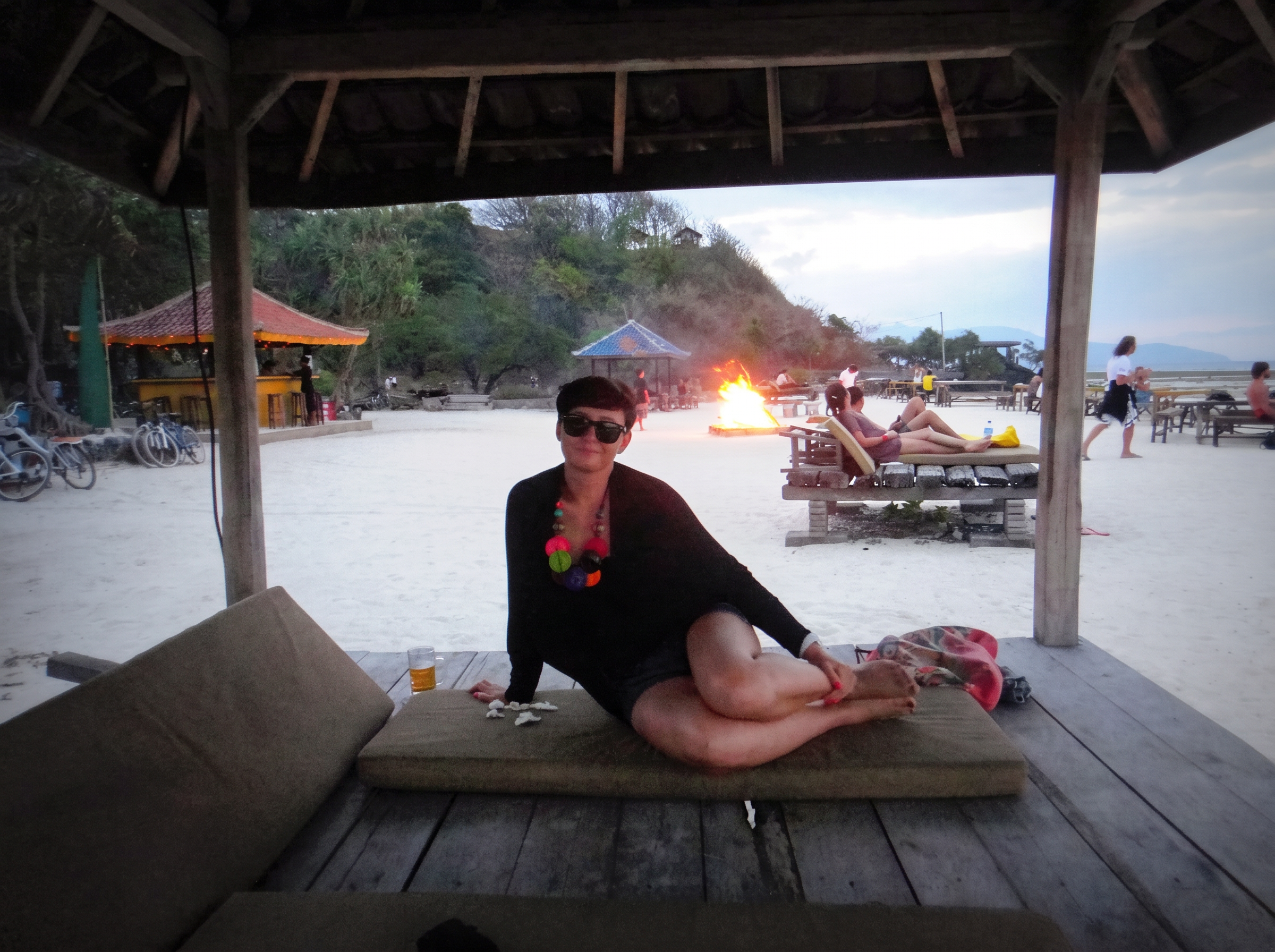 A woman relaxing on a beach lounge at dusk with a bonfire glowing on Gili Trawangan